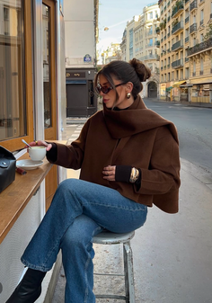 Woman in a brown jacket and blue jeans sitting outdoors on a street