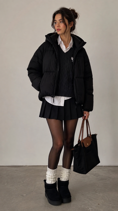 Woman in a black puffer jacket, pleated skirt, and winter boots holding a bag