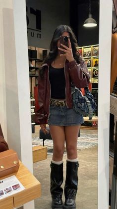 Woman in a denim skirt and leather jacket taking a mirror selfie indoors