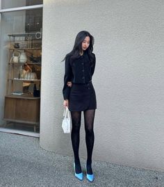 Woman in a black skirt suit and blue heels holding a white handbag outdoors