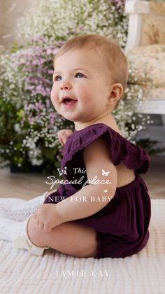 Happy baby in a purple romper sitting on a white blanket with flowers in the background