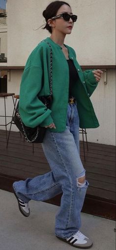 Woman in a green jacket, ripped jeans, and sunglasses walking outdoors