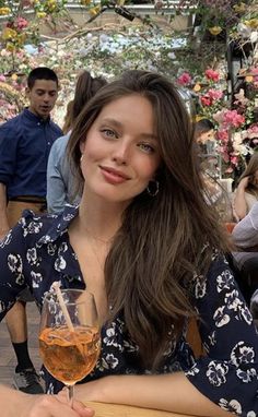 Woman with long brown hair in a floral dress holding a drink indoors