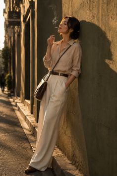 Woman in a loose shirt and wide leg pants leaning against a wall at golden hour