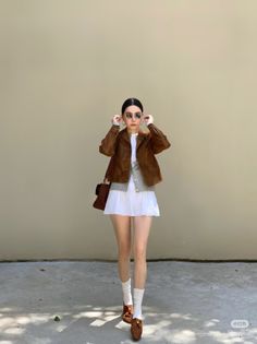 Woman in a brown jacket and white skirt adjusting her sunglasses outdoors