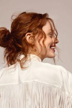 Woman with red hair in a messy bun, smiling in profile, wearing a white shirt with fringe