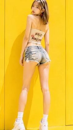 Woman in denim shorts and a strapless top posing against a yellow wall