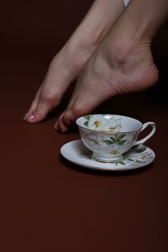 Bare feet positioned above a porcelain teacup with a floral pattern on a brown background