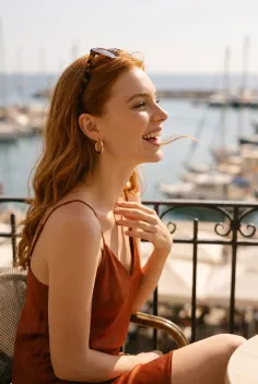 Smiling woman with red hair and sunglasses on her head, sitting outdoors overlooking a marina