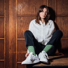 Woman in a white shirt and black jeans with patterned socks sitting against a wooden wall