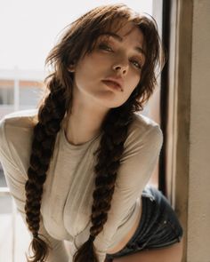 Woman with braids in a long sleeve shirt and denim shorts posing by a window