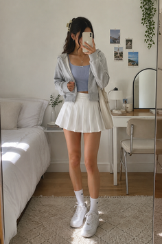Woman in a white pleated skirt and grey cropped hoodie taking a mirror selfie