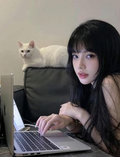Woman with long black hair using a laptop, with a white cat behind her