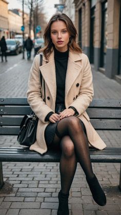 Woman in a beige coat and black tights sitting on a bench on a city street