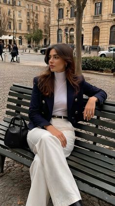 Woman with long hair wearing a navy blazer and white pants sitting on a bench outdoors