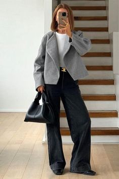 Woman in a gray jacket and wide leg pants holding a black handbag indoors