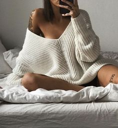 Woman in an oversized white sweater sitting on a bed, taking a selfie