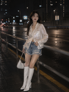 Woman in a sheer top, denim shorts, and white boots on a wet city street at night