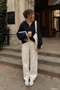 Woman in a navy sweater and white pants holding books and coffee on a campus
