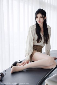 Woman with long black hair wearing a white shirt and brown shorts, sitting indoors