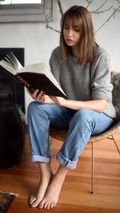 Woman with bangs reading a book while sitting barefoot in a chair