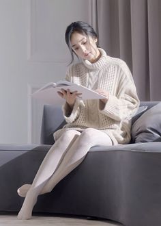 Woman in a cream turtleneck sweater and white tights reading a book on a sofa