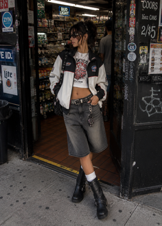 Woman in a crop top, racing jacket, and denim shorts standing outside a store
