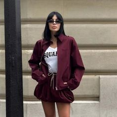 Woman with dark hair and sunglasses wearing a maroon jacket and shorts outdoors