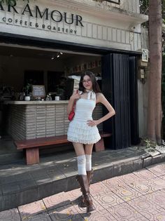 Woman in a white dress, brown boots, and glasses standing outside a coffee shop