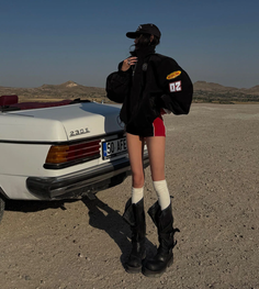Person in a racing jacket and red shorts standing next to a white convertible car