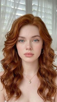 Woman with long red curly hair and blue eyes wearing a heart necklace indoors