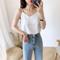 Woman in a white camisole and blue jeans taking a mirror selfie indoors