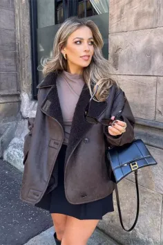 Woman in a brown shearling jacket and black mini skirt holding sunglasses