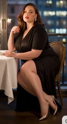 Woman in a black dress and high heels sitting at a table with a city view at night
