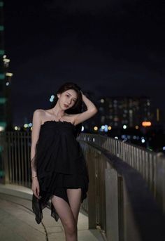 Woman in a black dress posing on a bridge at night with city lights in background