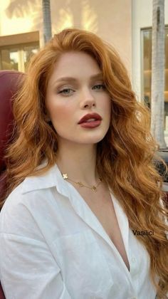 Woman with long red hair and red lipstick wearing a white shirt and gold necklace
