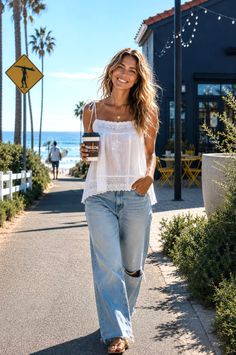 Woman with long hair in a white top and ripped jeans walking outdoors near a beach