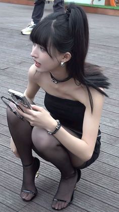 Woman in a black tube top and pantyhose squatting outdoors while holding a phone