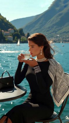 Woman in a black dress and pearls drinking coffee by a lake with mountains