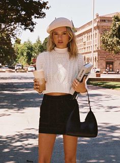 Woman in a beret and cropped sweater holding a drink and books on a sunny street