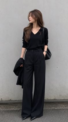 Woman in a black top and wide leg pants standing against a gray wall