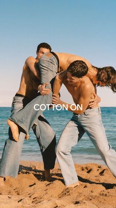 Three shirtless men in jeans on a sandy beach with the ocean in the background