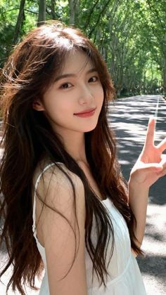 Woman with long hair in a white top making a peace sign on a tree-lined road