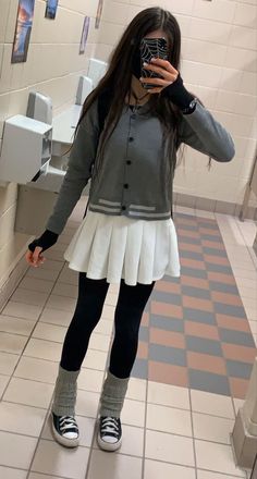 Person in a pleated skirt, cardigan, and leg warmers taking a mirror selfie indoors