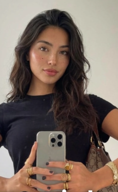 Woman with long wavy hair and freckles holding an iPhone, wearing a black t-shirt and gold rings
