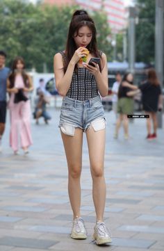Woman in denim shorts and a patterned top drinking and looking at her phone on a street