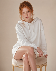 Woman with red hair and freckles wearing a white hoodie, sitting on a stool