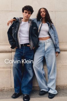 Two people in denim jackets and loose jeans leaning against a wall