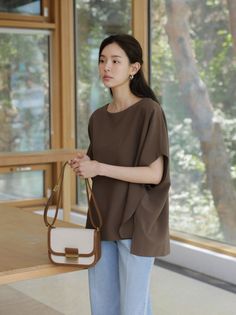 Woman in a brown top and light blue pants holding a shoulder bag indoors