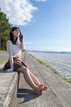 Woman in a white lace top and brown skirt sitting barefoot on a concrete ledge by a beach